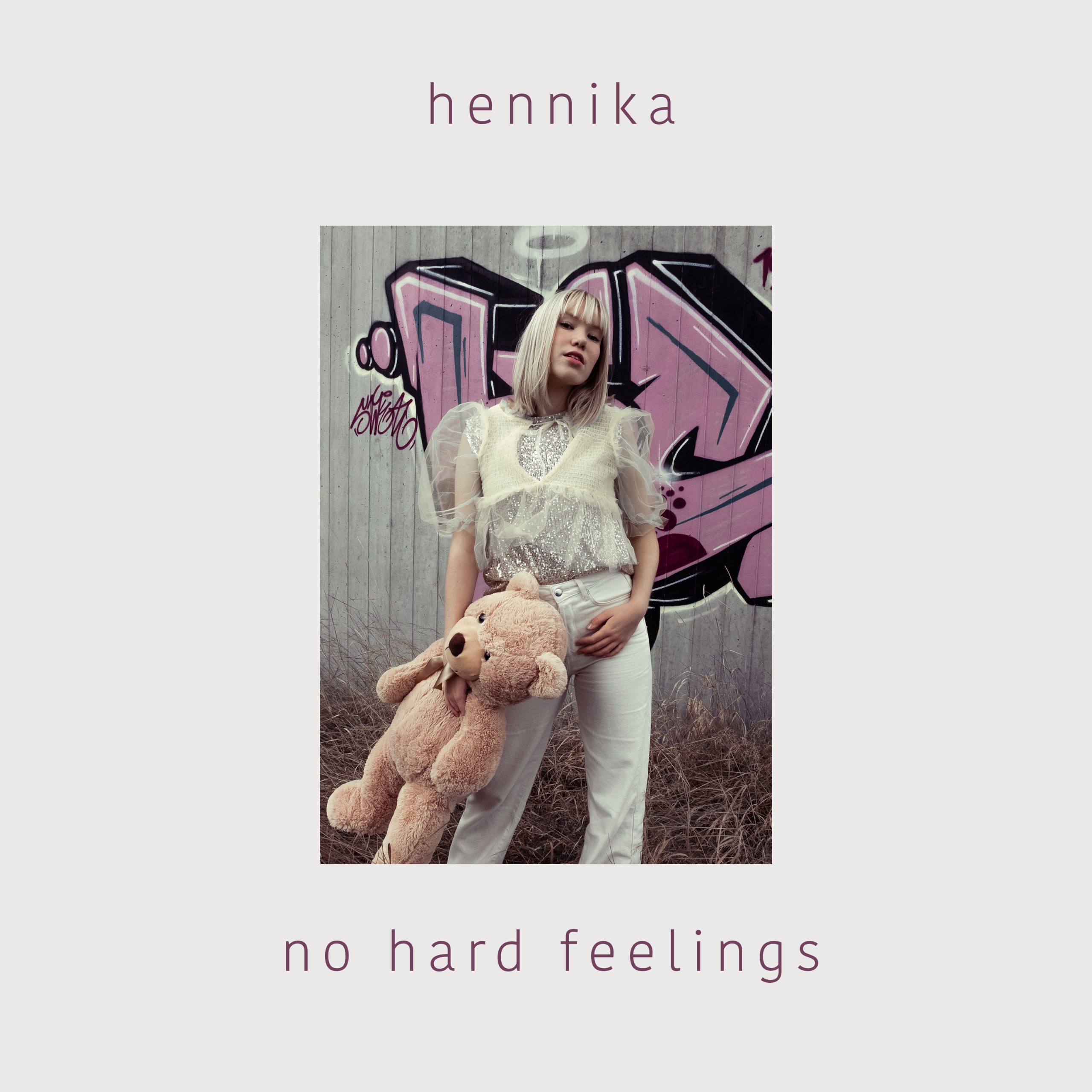 Viser coveret til Hennika sin nyeste låt No hard feelings. Hun står foran grafitti og holder en stor teddybjørn i hånden.